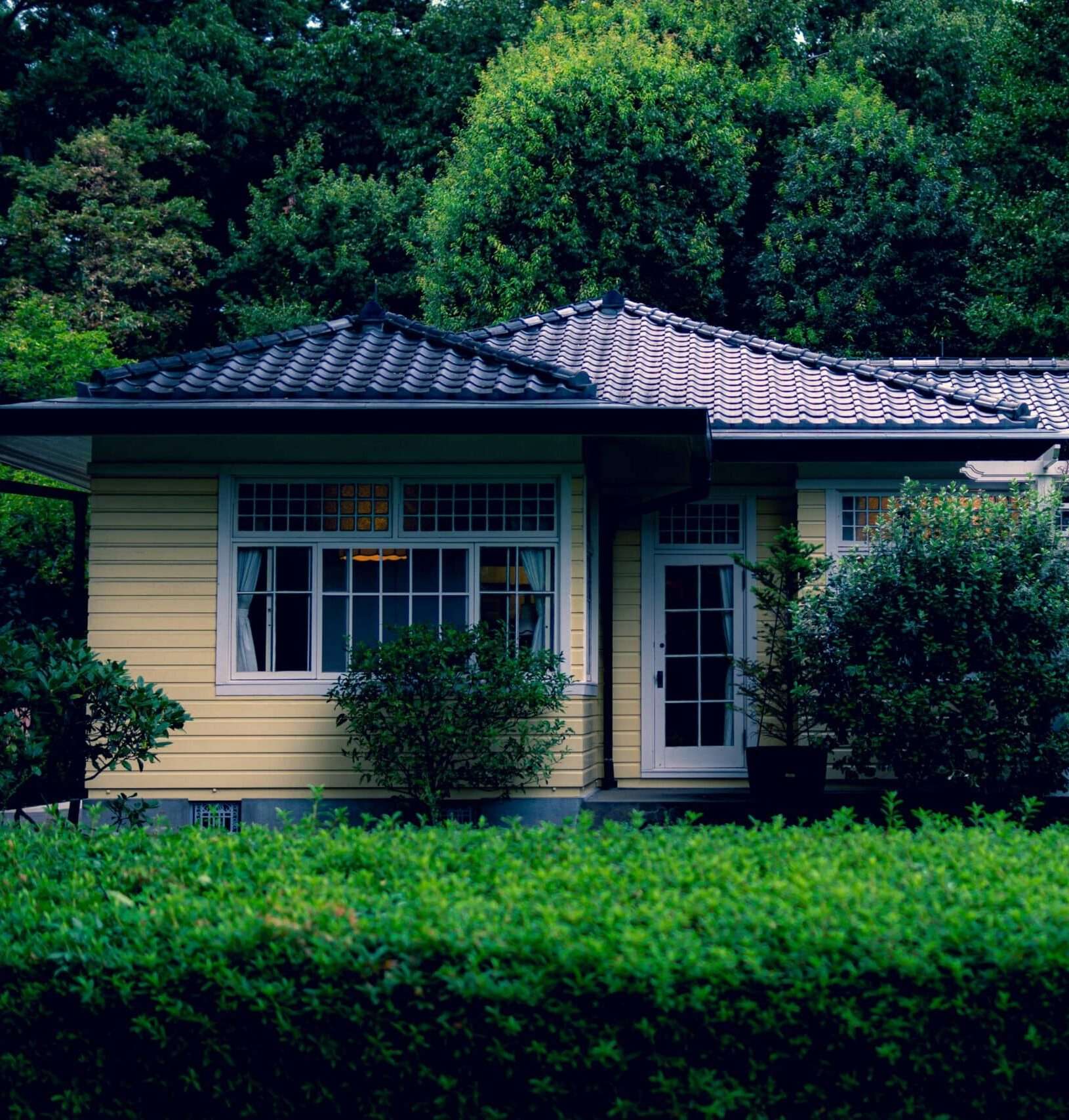 Small yellow home with tiled roof surrounded by trees and greenery in a quiet residential setting